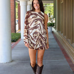 A woman modeling a one-shoulder mini dress with a brown and beige tiger print, paired with dark brown cowboy boots, walking along a covered outdoor walkway near Rodéo Boutique.
