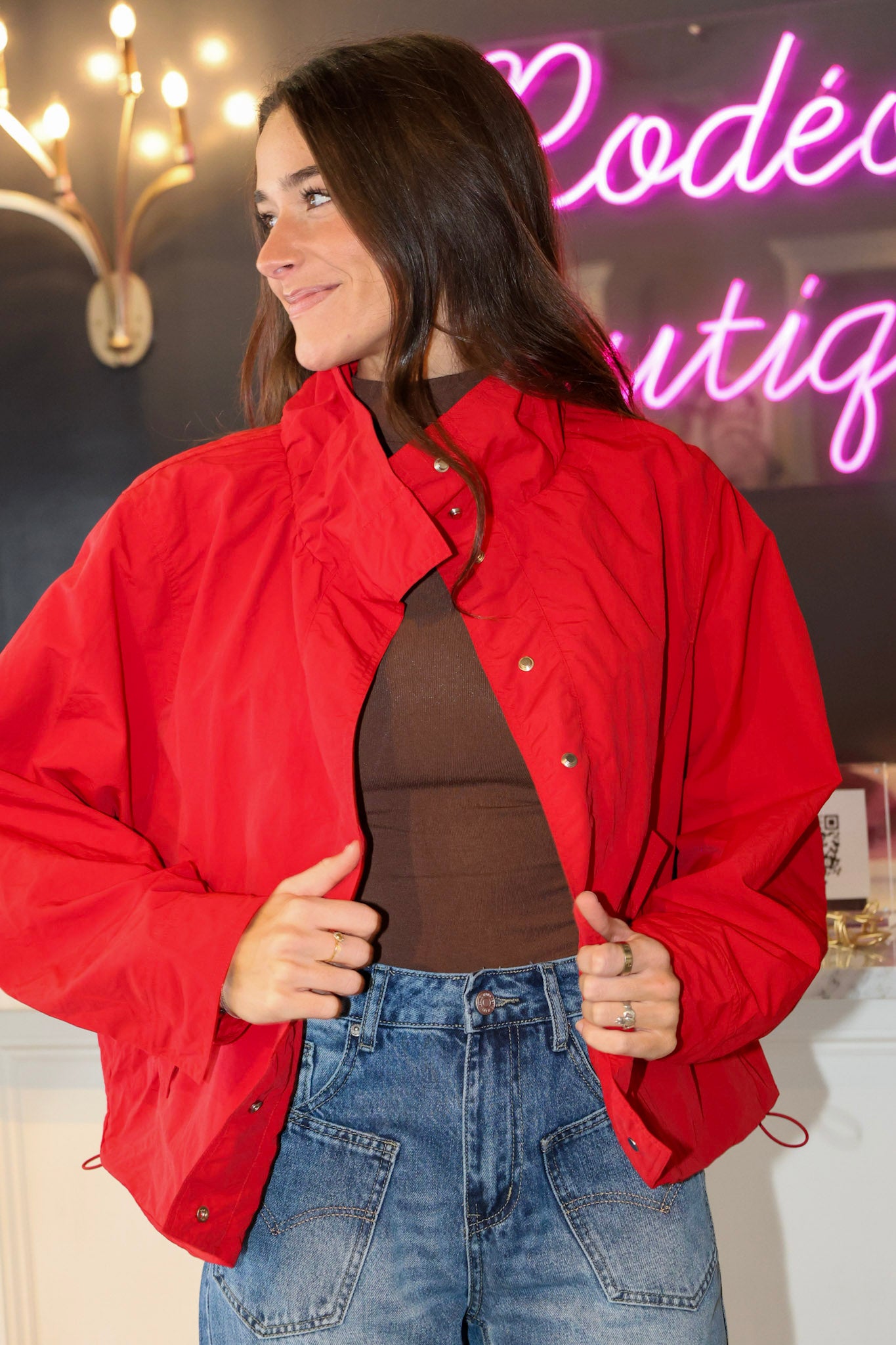 A woman wearing a bright red cropped jacket over a brown top and blue jeans, standing in a boutique setting with a pink neon sign in the background.