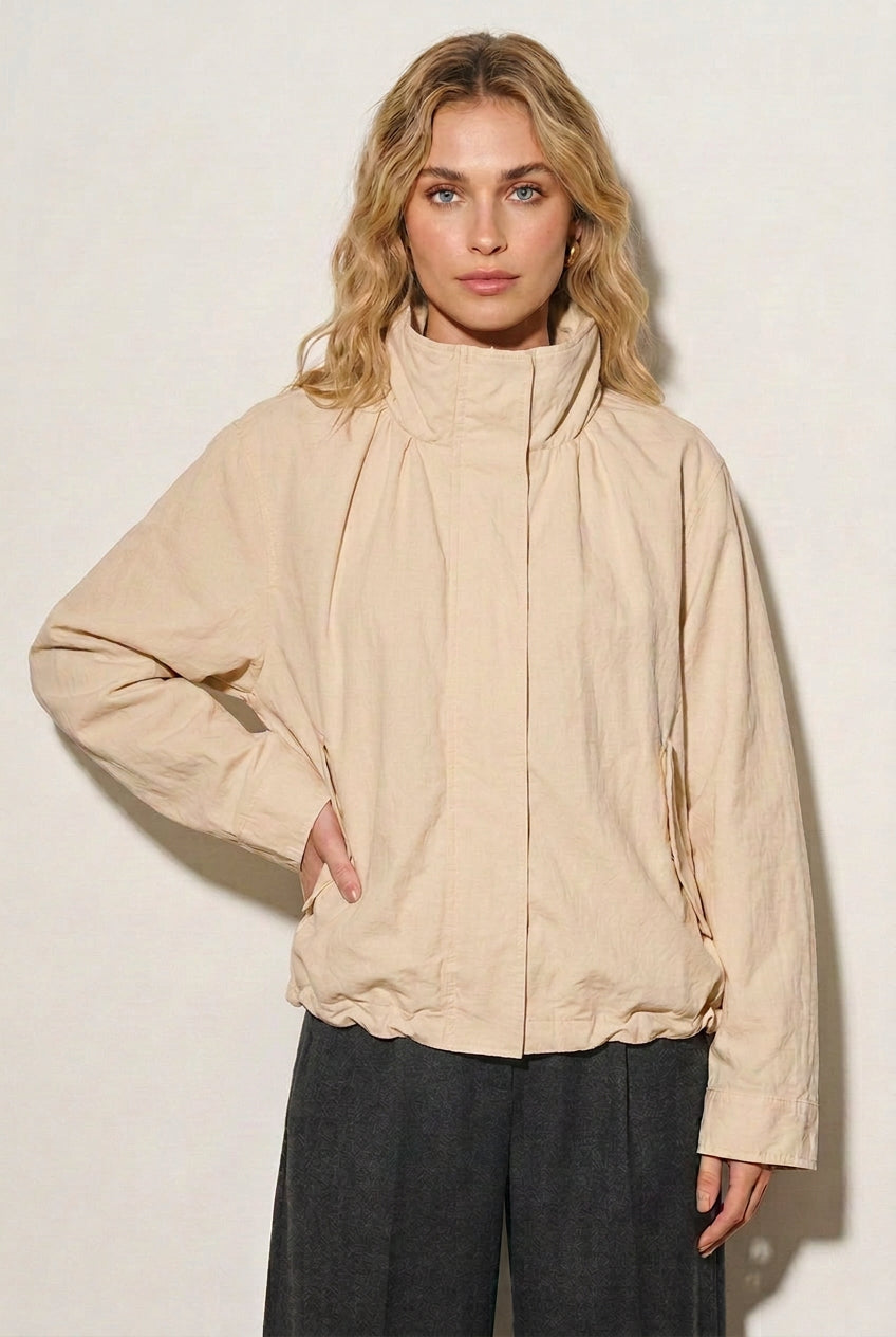 A woman modeling an oversized cream-colored lightweight jacket with a high collar and front zipper, paired with dark gray pants, against a plain off-white background.