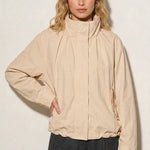 A woman modeling an oversized cream-colored lightweight jacket with a high collar and front zipper, paired with dark gray pants, against a plain off-white background.