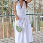 Woman in a white dress holding a green straw bag on a wooden deck with trees in the background