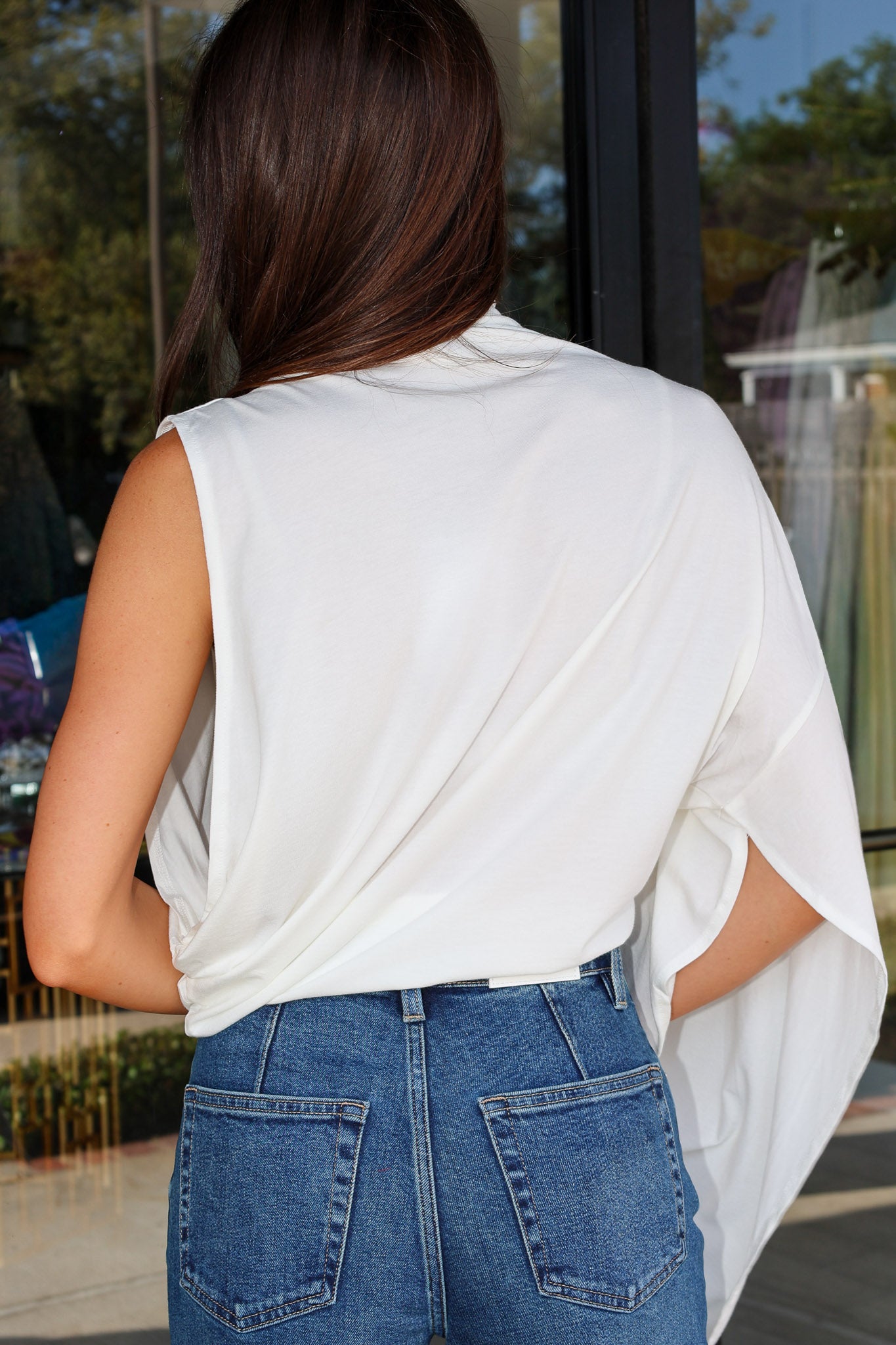 A woman viewed from behind wearing a sleeveless white draped top and blue jeans in front of a storefront window.