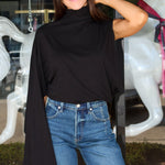 A woman wearing a black draped cape-sleeve mock-neck top and blue jeans, standing in front of a glass storefront with white horse statues visible in the background.
