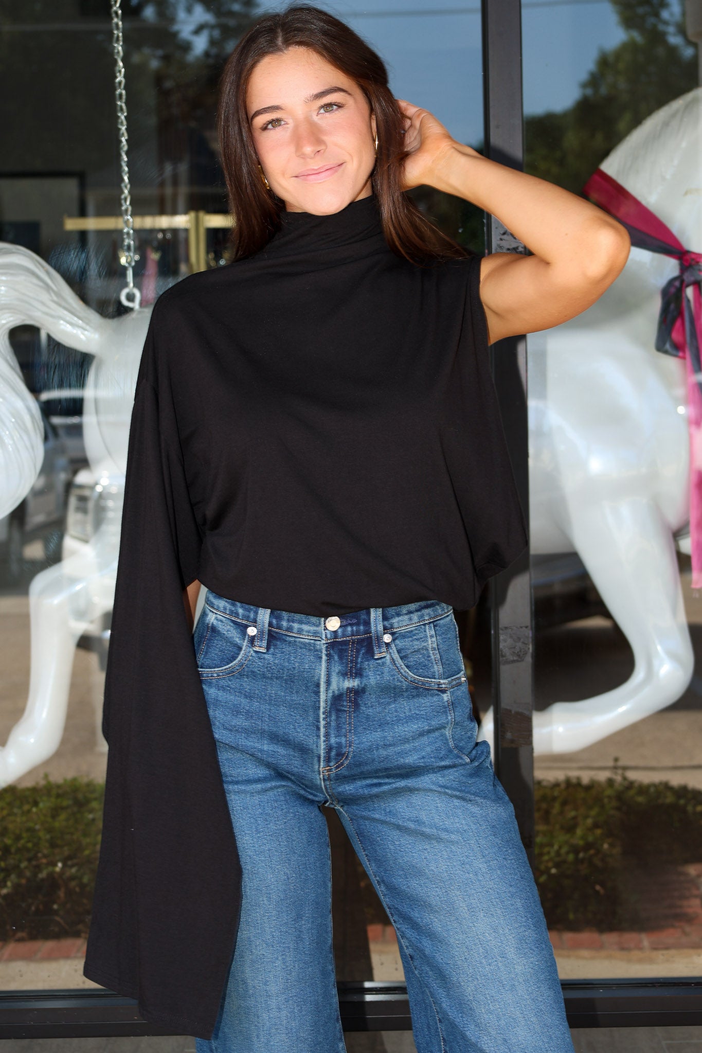 A woman wearing a black draped cape-sleeve mock-neck top and blue jeans, standing in front of a glass storefront with white horse statues visible in the background.
