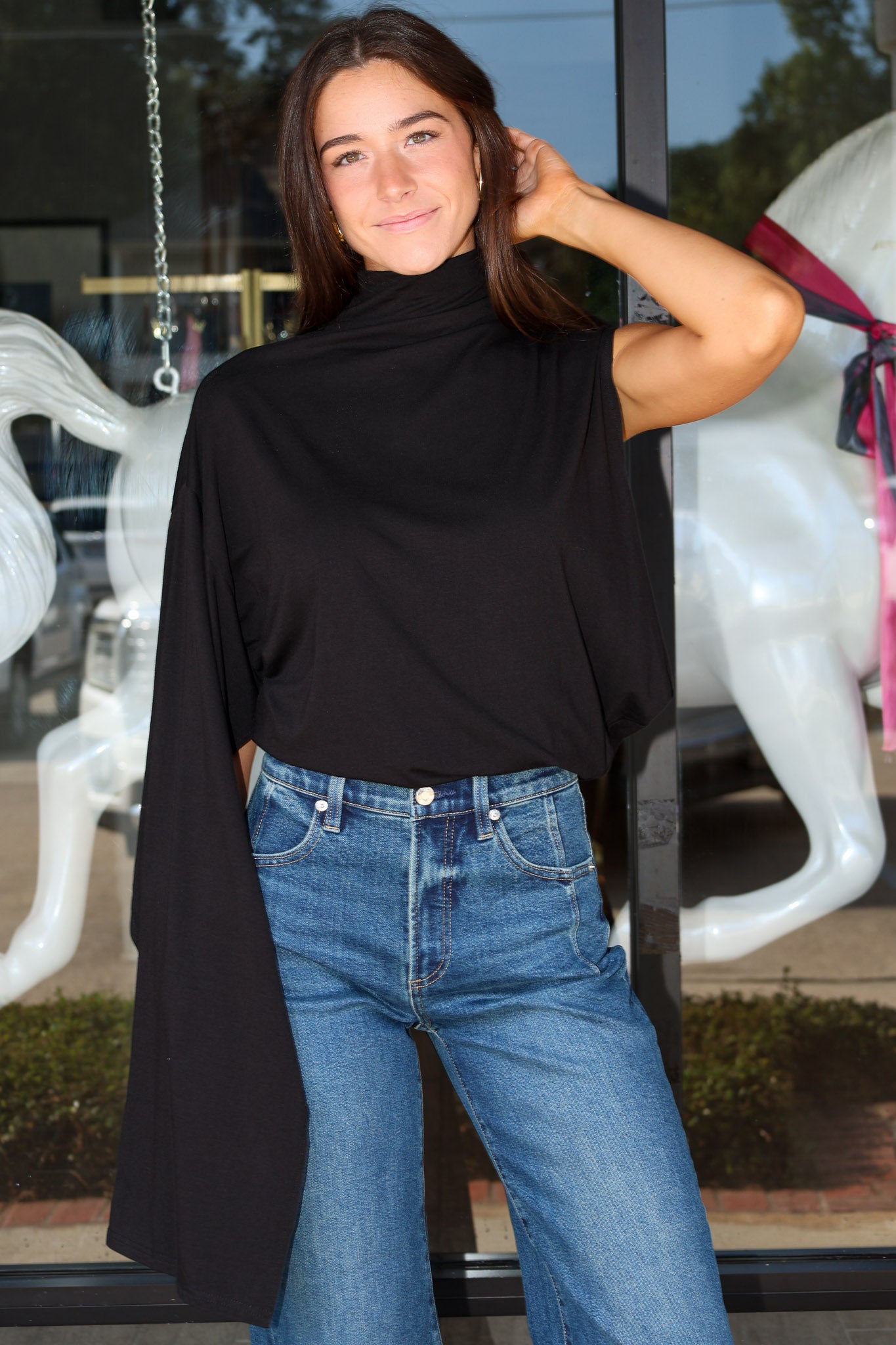 A woman wearing a black draped cape-sleeve mock-neck top and blue jeans, standing in front of a glass storefront with white horse statues visible in the background.