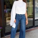 A woman stands in front of a glass storefront wearing a white asymmetrical draped top, wide-leg blue jeans, and bright pink sneakers.