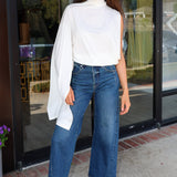 A woman stands in front of a glass storefront wearing a white asymmetrical draped top, wide-leg blue jeans, and bright pink sneakers.