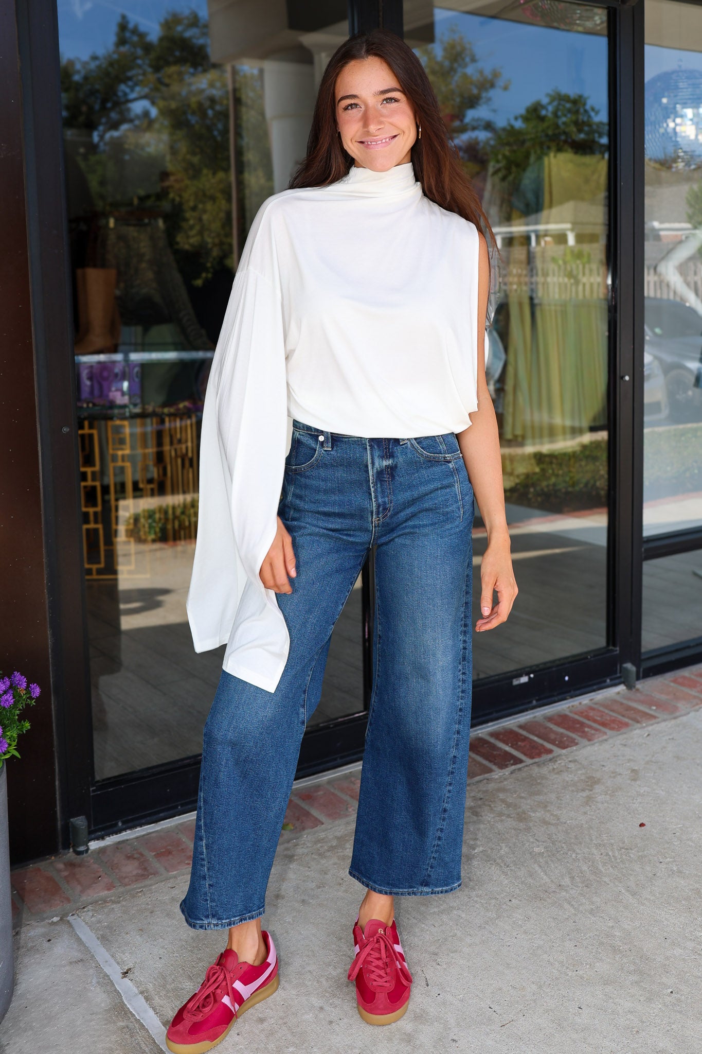 A woman stands in front of a glass storefront wearing a white asymmetrical draped top, wide-leg blue jeans, and bright pink sneakers.