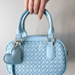 Light blue woven handbag with a heart-shaped tag held by a hand against a neutral background
