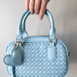 Light blue woven handbag with a heart-shaped tag held by a hand against a neutral background