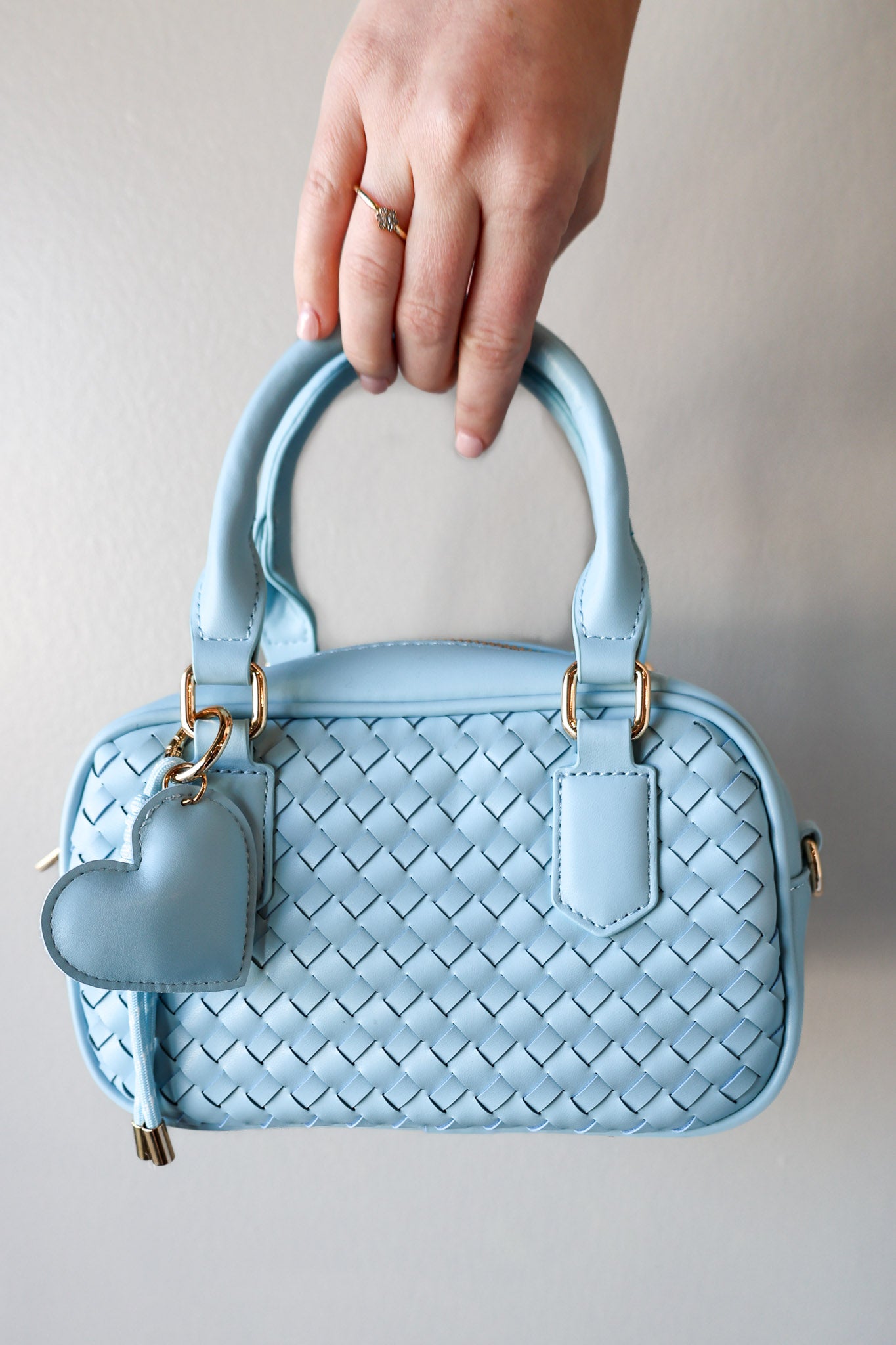 Light blue woven handbag with a heart-shaped tag held by a hand against a neutral background