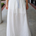 Person wearing a long white skirt with eyelet details on a sidewalk.