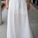 Person wearing a long white skirt with eyelet details on a sidewalk.