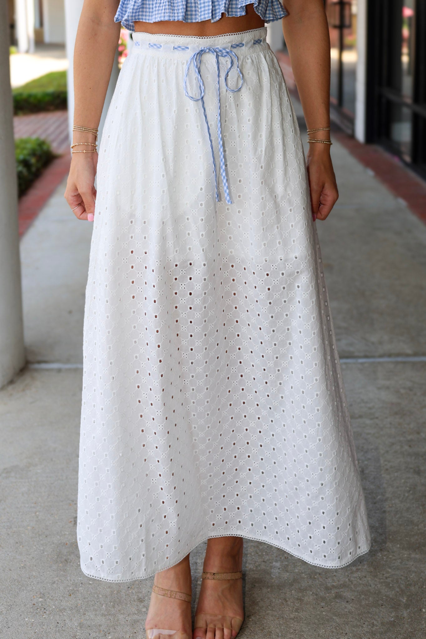 Person wearing a long white skirt with eyelet details on a sidewalk.