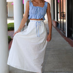 Woman wearing a blue top and white skirt standing on a sidewalk.