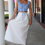Woman wearing a blue top and white skirt standing on a sidewalk.